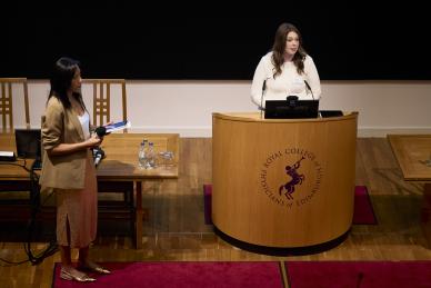 Woman stands on left looking at woman speaking at podium